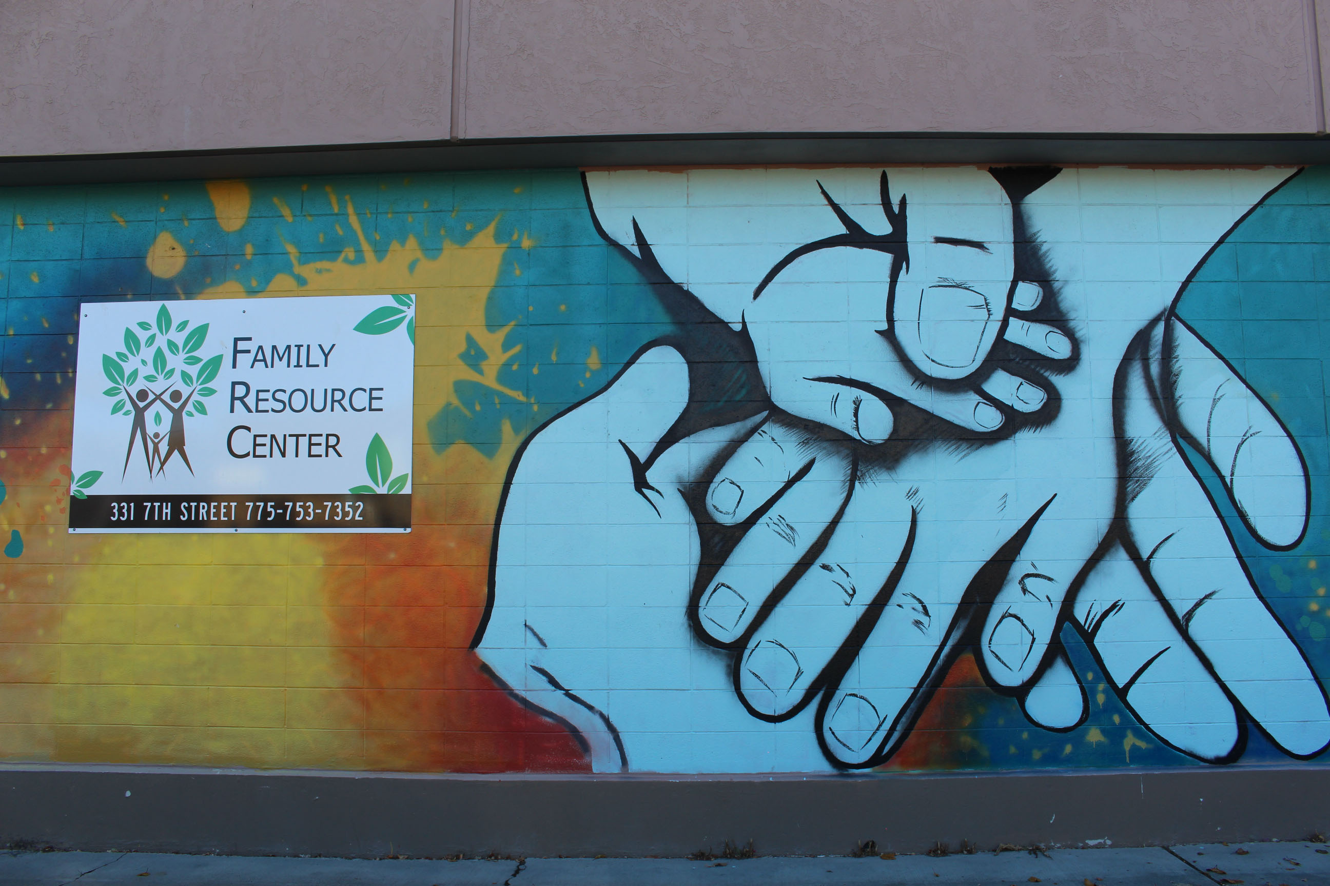 A large mural has been painted on the side of a building. It shows hands of different sizes and ages holding each other over a rainbow background. On the left, a sign reads: Family Resource Center